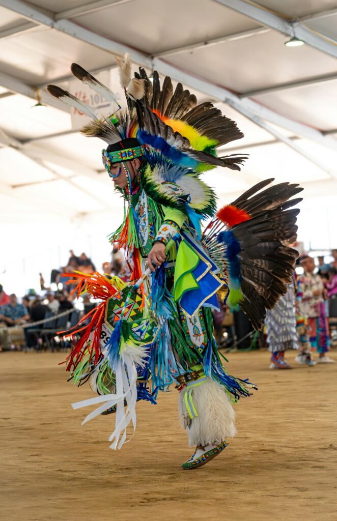 male dancer in colourful regalia, caught in dance movement between a metal tent structure - Photo by Soly Moses: https://www.pexels.com/photo/vibrant-native-american-pow-wow-dancer-in-traditional-dress-28820284/