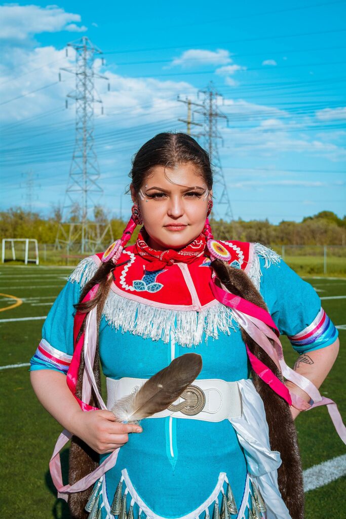 indigenous woman in sky blue regalia with red and white ribbons and fringe, holds and feather and stands proudly in a field - Photo by Farouk Animashaun: https://www.pexels.com/photo/portrait-of-a-woman-in-traditional-clothes-12620918/
